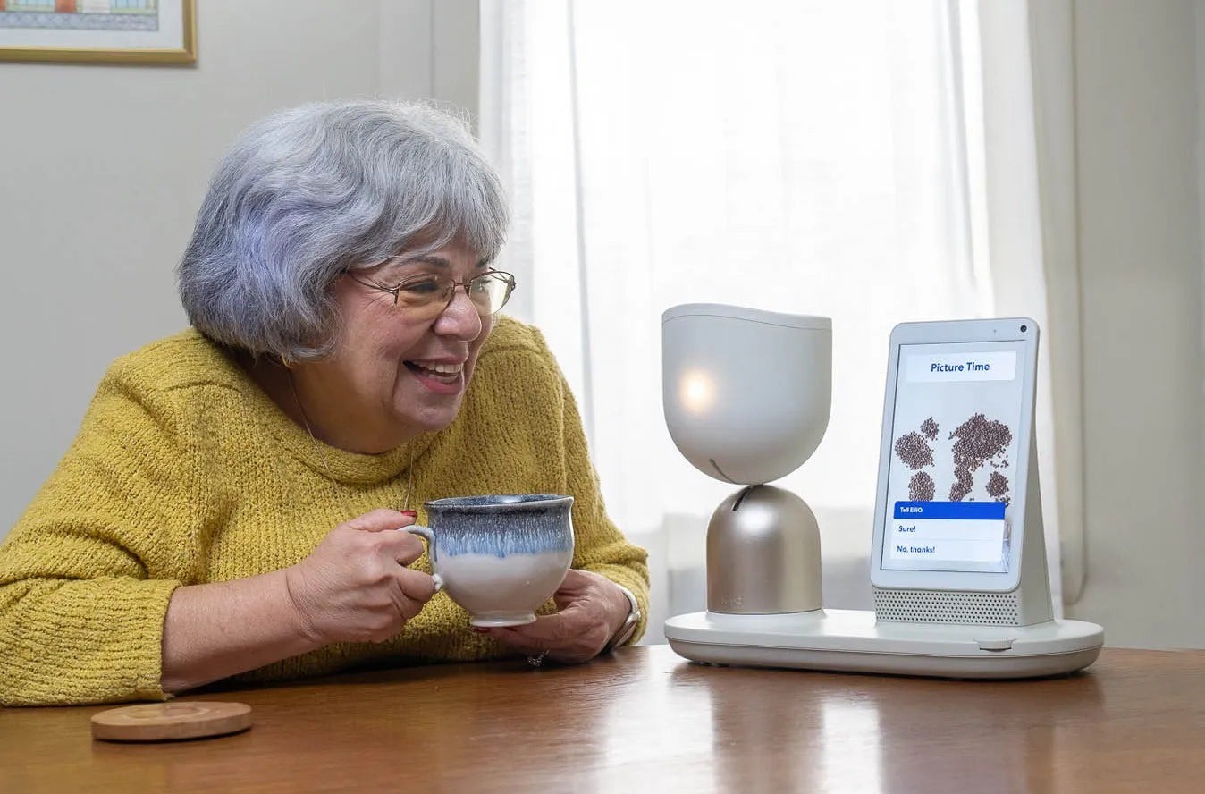 An older woman with a coffee cup laughs next to ElliQ, a small tabletop robot with a screen